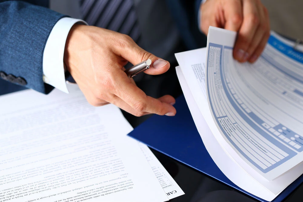 Male arm in suit offer insurance form clipped to pad and silver pen to sign closeup.