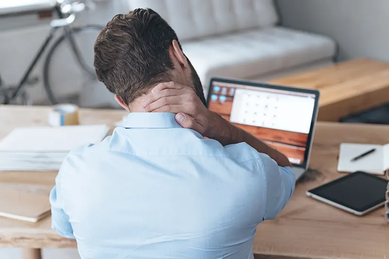 Rear view of frustrated young man looking exhausted and massaging his neck while sitting at his working place. Occupational disease.